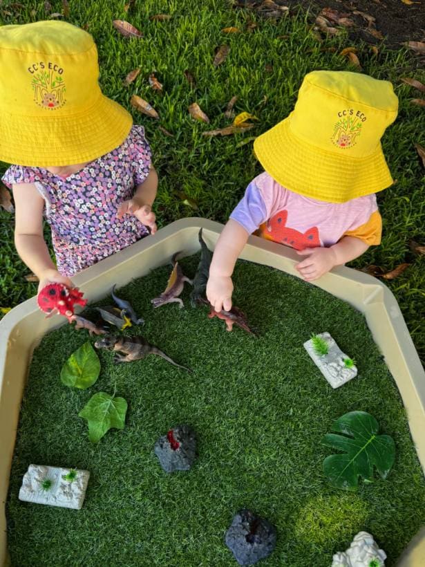 Duas crianças de chapéu explorando juntas uma bandeja sensorial no jardim