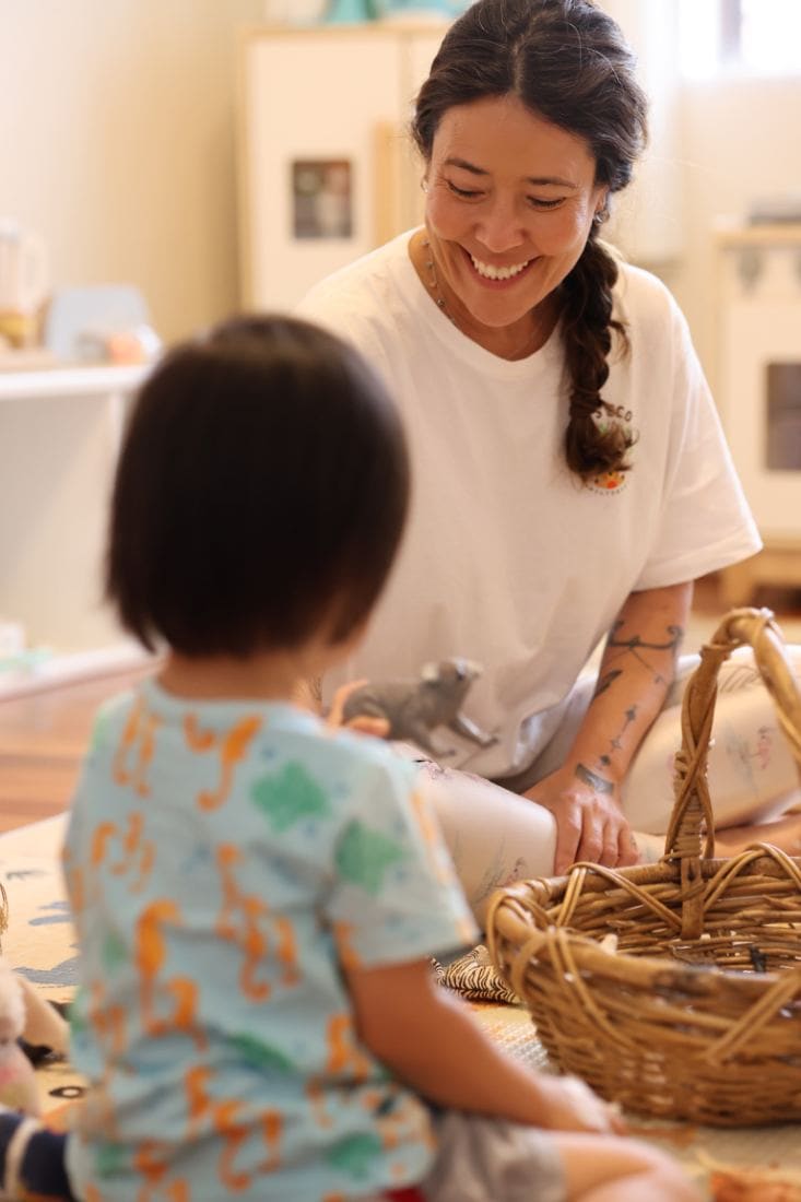 Cintia B Lemm sorrindo enquanto brinca com uma criança em uma cesta de materiais naturais